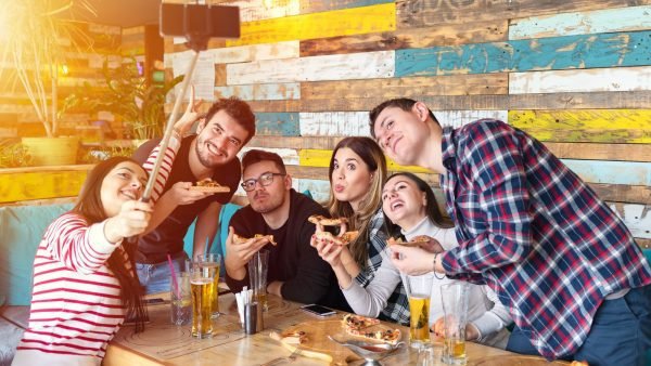 Cheerful young friends taking selfie with smart phone while sharing a pizza, Young people at table in restaurant using mobile phone to take selfie while eating and share on social media