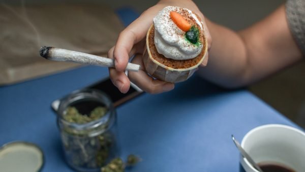 Young woman smoking a joint of marijuana while eats coffee and cake for breakfast. Glass jar full of cannabis buds on the table, blur effect.